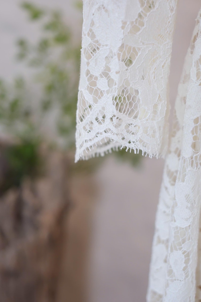 May include: An off-white lace garment featuring long sleeves with intricate floral patterns and scalloped edges. The delicate fabric displays a detailed openwork design throughout, highlighting the craftsmanship of the lace texture and the hem.