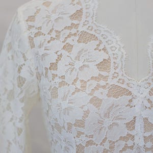 May include: A long-sleeved top made of white floral lace featuring a scalloped V-neckline. The intricate lace pattern covers the entire garment, showcasing a detailed botanical design with a delicate mesh background and finished edges.
