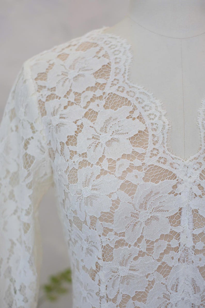 May include: A long-sleeved top made of white floral lace featuring a scalloped V-neckline. The intricate lace pattern covers the entire garment, showcasing a detailed botanical design with a delicate mesh background and finished edges.