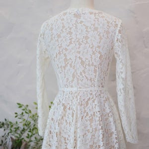 May include: A long-sleeved dress featuring an intricate white floral lace overlay atop a nude-colored lining. The garment has a fitted bodice, a defined waistline, and a flared skirt, displayed on a mannequin to show the back view.