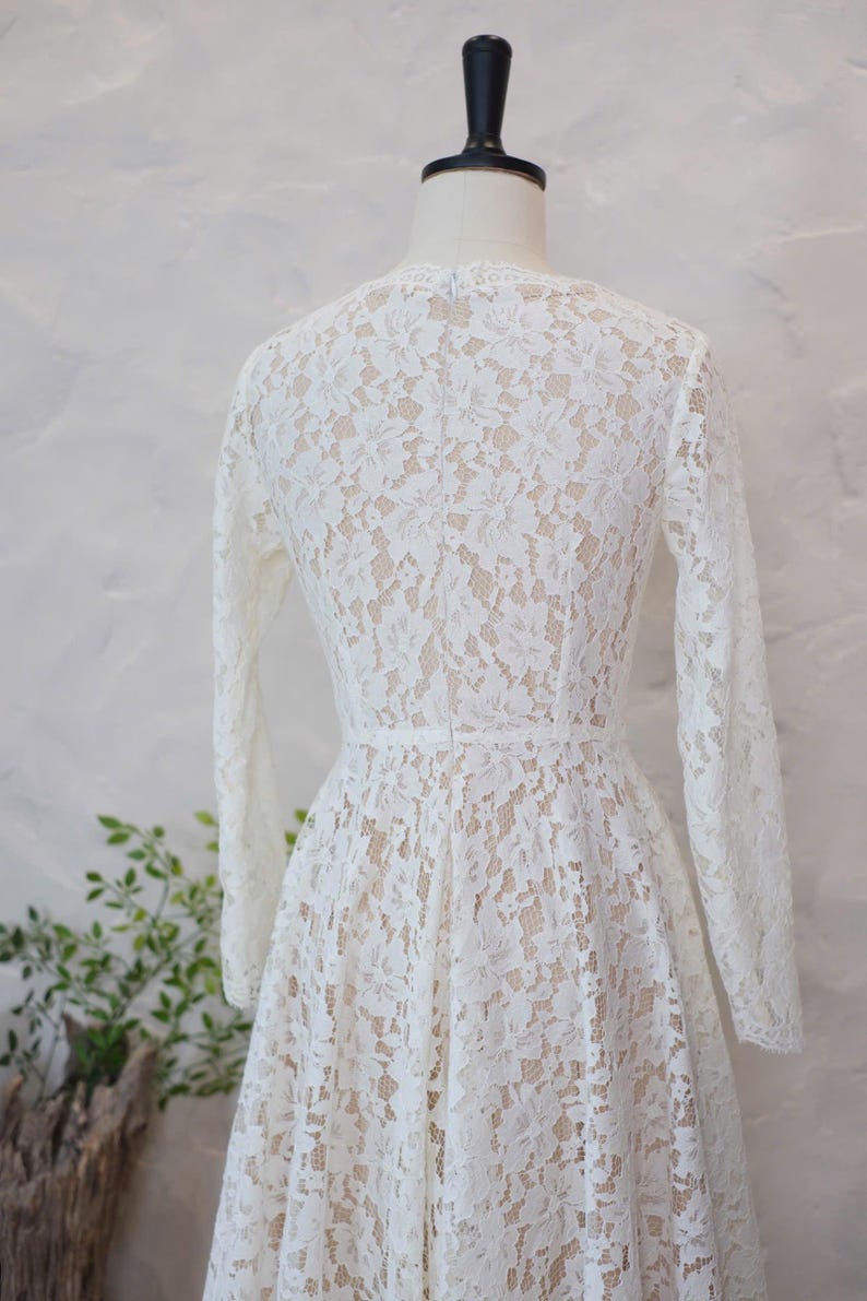 May include: A long-sleeved dress featuring an intricate white floral lace overlay atop a nude-colored lining. The garment has a fitted bodice, a defined waistline, and a flared skirt, displayed on a mannequin to show the back view.