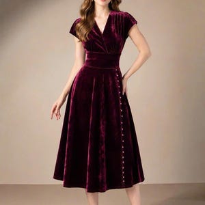 Puede incluir: Un vestido midi de terciopelo color burdeos intenso con escote en V, mangas cortas y cintura entallada. La falda tiene una silueta acampanada con una hilera vertical de pequeños botones claros que bajan por el lado izquierdo desde la cintura hasta el dobladillo.