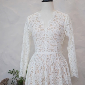 May include: A long-sleeved dress featuring white floral lace overlaying a nude-colored lining. The garment has a scalloped V-neckline, a fitted bodice, and a flared skirt. The lace pattern covers the entire dress, including the sleeves.