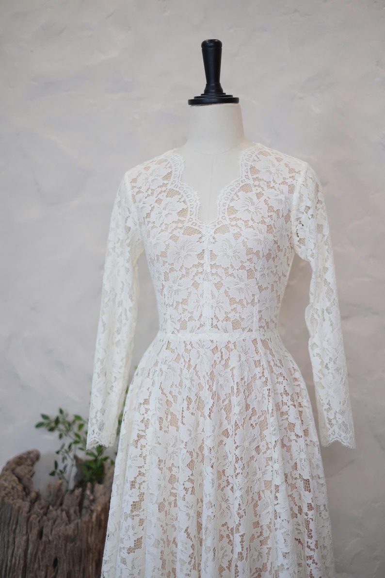 May include: A long-sleeved dress featuring white floral lace overlaying a nude-colored lining. The garment has a scalloped V-neckline, a fitted bodice, and a flared skirt. The lace pattern covers the entire dress, including the sleeves.