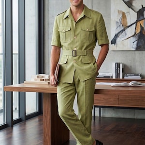 Op de afbeelding: Een licht olijfgroene safari-set bestaande uit een shirt met korte mouwen, een bijpassende riem en een broek. Het shirt heeft een puntige kraag, epauletten op de schouders en vier voorzakken met knoopsluitingen. De broek heeft rechte pijpen.