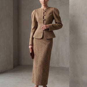 Peut inclure: Un ensemble deux pièces beige composé d'une veste à manches longues avec une encolure ronde, quatre boutons sur le devant et des épaules bouffantes, assorti d'une jupe longue jusqu'aux chevilles. Le tissu présente un motif tissé en diagonale.