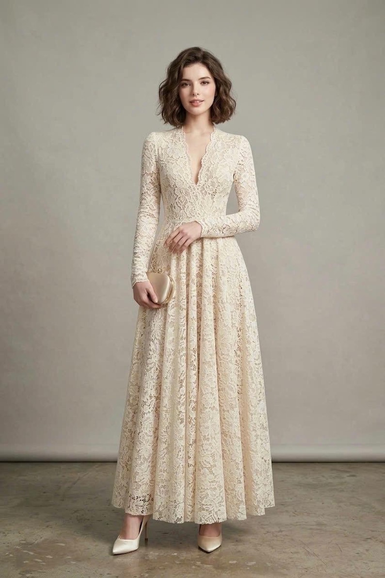 May include: A floor-length cream-colored lace dress featuring long sleeves, a deep V-neckline, and a fitted bodice that transitions into a full, flowing skirt. The garment is paired with a small, smooth, metallic gold-toned clutch and pointed-toe heels.