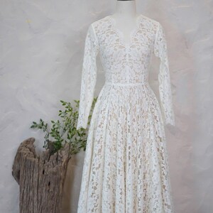 May include: A white lace dress with long sleeves, a V-neckline, and a fitted bodice that transitions into a full, pleated skirt. The dress is displayed on a mannequin and features the text A version with a higher neckline at the top.