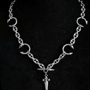 Pode incluir: Colar prateado com design de corrente, detalhes pontiagudos e um pingente de adaga. O colar tem uma estética gótica ou punk, com um estilo escuro e ousado. A corrente é feita de metal e tem um acabamento polido.