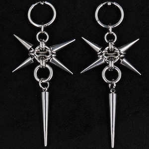 May include: A pair of silver-toned metal earrings featuring a circular hoop at the top, connected to a central ring with four radiating spikes, and a single long, pointed spike dangling from the bottom of each earring assembly.