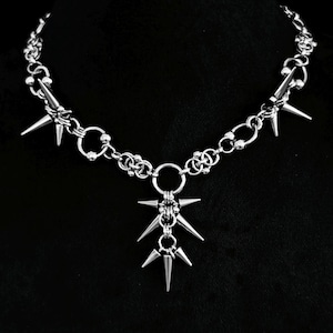 Könnte beinhalten: Eine silberfarbene Kettenhalskette mit einer Reihe von miteinander verbundenen Metallringen und kleinen kugelförmigen Perlen. Das Design umfasst mehrere scharfe, konische Stacheln, die an den Kettengliedern befestigt sind, mit einem zentralen Bündel aus drei herabhängenden Stacheln.