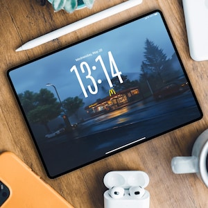 Op de afbeelding: Een digitale tablet toont een nachtscène met een McDonald's restaurant. Een witte stylus, een oranje telefoon en witte draadloze oortelefoons zijn rond de tablet op een houten oppervlak geplaatst. Een kleine vetplant in een blauwe pot is ook aanwezig.