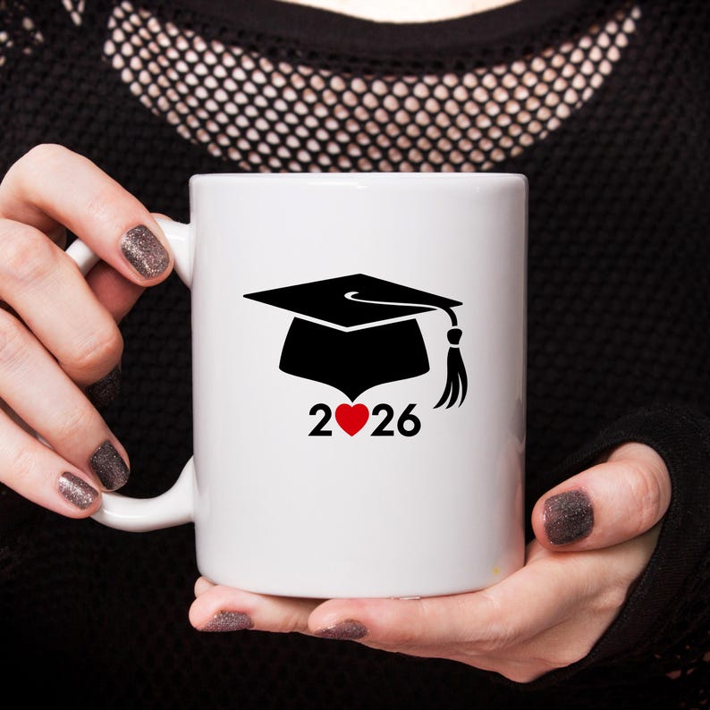 May include: White ceramic mug featuring a black graduation cap design with a tassel and the year 2026. A red heart is between the numbers. The mug is held by a person with dark nail polish.