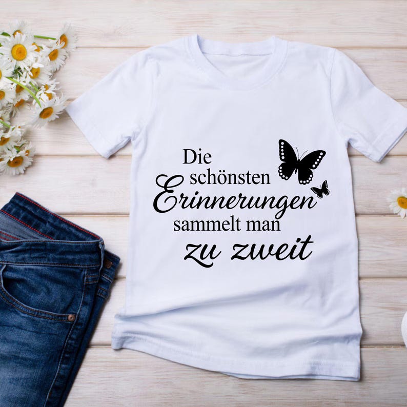 May include: A white short-sleeved t-shirt featuring black text that reads Die sch&ouml;nsten Erinnerungen sammelt man zu zweit, accompanied by two black butterfly silhouettes. The shirt is displayed alongside blue denim jeans and white flowers.
