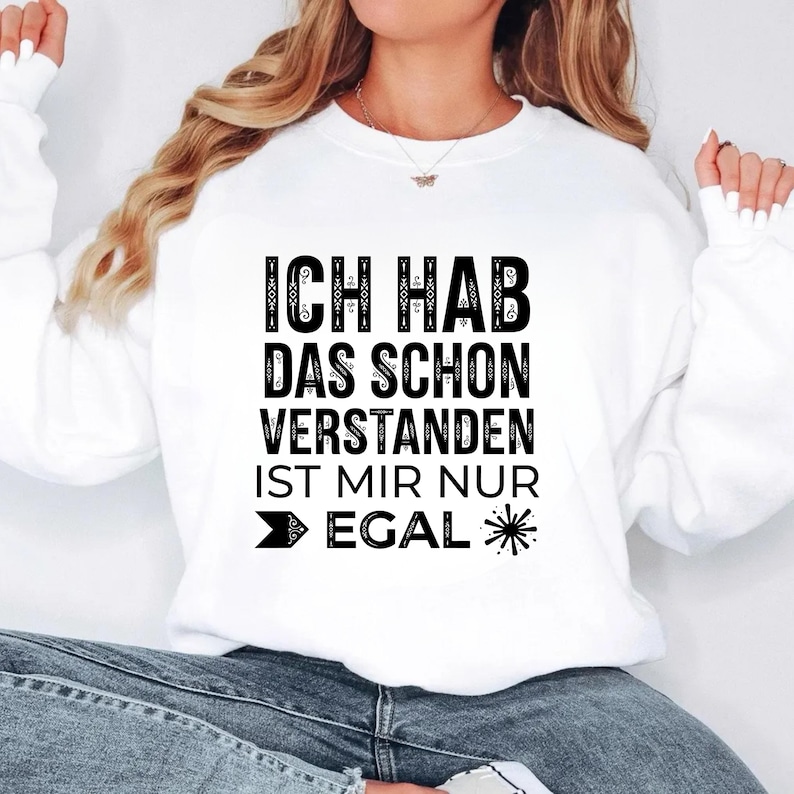 May include: A white long-sleeve sweatshirt featuring black text that reads "ICH HAB DAS SCHON VERSTANDEN IST MIR NUR EGAL" with a small arrow icon and a starburst graphic. The garment has a crew neckline and ribbed cuffs on the sleeves.