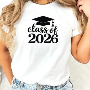 May include: White t-shirt featuring a black graduation cap and the words "class of 2026". The design is centered on the front of the shirt. The shirt is made of a soft material.