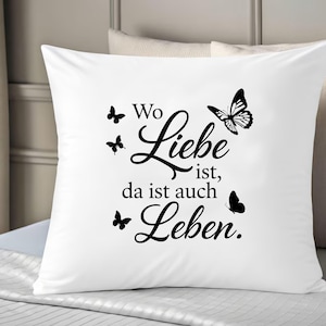 May include: A square white throw pillow featuring black text that reads Wo Liebe ist, da ist auch Leben. The design includes four black butterfly silhouettes of varying sizes arranged around the German script, creating a contrast on the fabric.