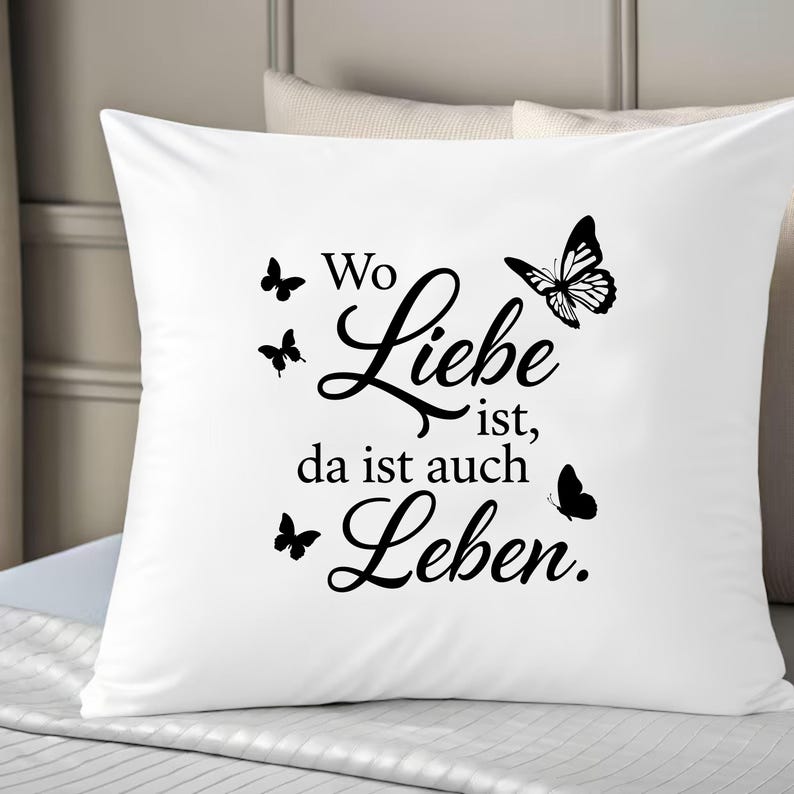 May include: A square white throw pillow featuring black text that reads Wo Liebe ist, da ist auch Leben. The design includes four black butterfly silhouettes of varying sizes arranged around the German script, creating a contrast on the fabric.