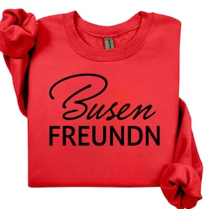 May include: A bright red long-sleeve sweatshirt featuring the black text Busen FREUNDN printed on the front. The garment has a crew neckline and ribbed cuffs on the sleeves, showcasing a bold colour contrast between the red fabric and black lettering.