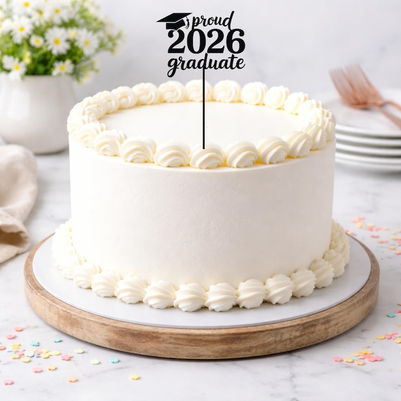 May include: A white frosted cake with decorative frosting around the top and bottom edges. A black cake topper reads "proud 2026 graduate" with a graduation cap. The cake sits on a wooden cake stand.