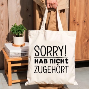 May include: A cream-coloured canvas tote bag featuring the black printed text SORRY! HAB nicht ZUGEH&Ouml;RT. The bag has two long shoulder straps and a rectangular shape, constructed from a woven fabric material with a clean, simple design.