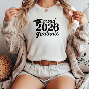 May include: Cream-colored t-shirt with the text "proud 2026 graduate" in black, featuring a graduation cap graphic. The shirt is paired with a brown belt and shorts. The person is wearing a beige cardigan.