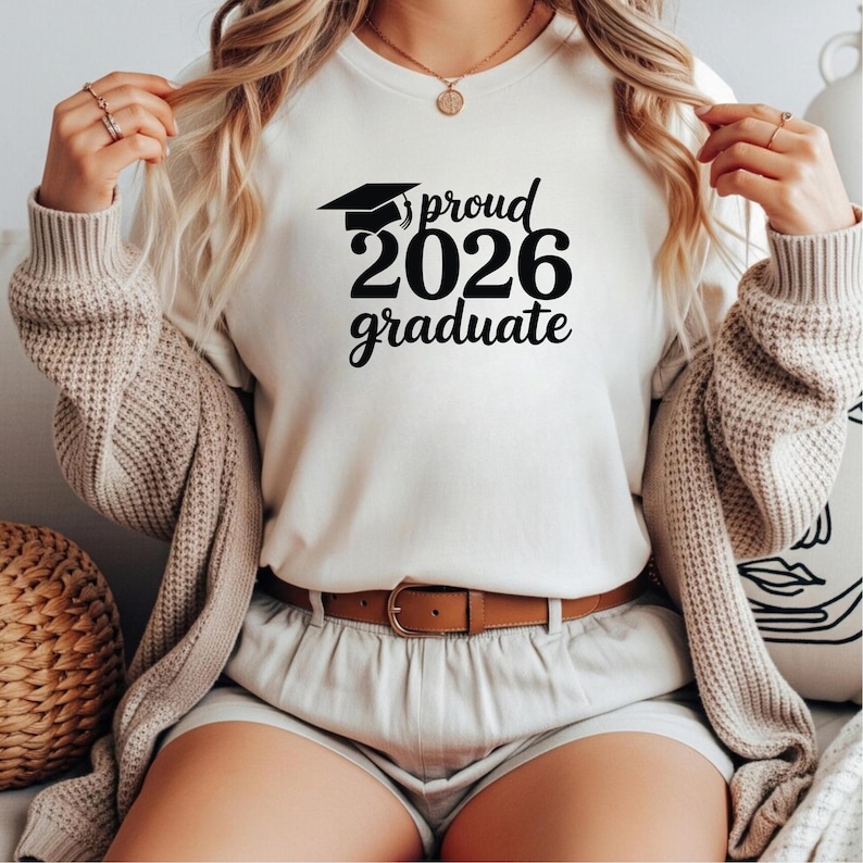 May include: Cream-colored t-shirt with the text "proud 2026 graduate" in black, featuring a graduation cap graphic. The shirt is paired with a brown belt and shorts. The person is wearing a beige cardigan.