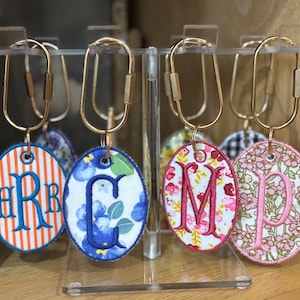 Puede incluir: Cuatro llaveros ovalados con mosquetones de metal dorado colgados en un soporte de acrílico transparente. Cada llavero presenta monogramas bordados sobre tela: rayas naranjas y blancas con letras azules, flores azules con letra azul, flores rosas con letra rosa y un patrón floral rosa con letra rosa.