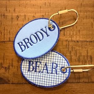 May include: Two oval-shaped fabric keychains with gold-toned metal loops. The top keychain is light blue with the name BRODY embroidered in dark blue. The bottom keychain features a light blue and white gingham pattern with BEAR embroidered in blue.