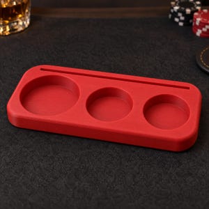 May include: A rectangular red tray with rounded corners featuring three circular recessed compartments of varying sizes and a long, narrow horizontal slot along the top edge, designed for holding small items or gaming accessories.