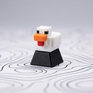 May include: A white and orange pixelated chicken keycap with a black base. The chicken has a red tongue and black eyes. The keycap is designed to fit on a mechanical keyboard.