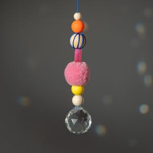 Könnte beinhalten: Ein dekorativer Hängeornament mit einem durchsichtigen Kristallkugel am Boden, mit einer Reihe farbiger Perlen und einem rosa Pompon. Die Perlen sind orange, gelb und weiß, mit einer blauen Schnur. Das Ornament hängt vor einem grauen Hintergrund.