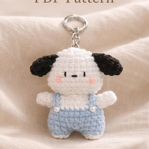 May include: A handmade crochet keychain featuring a white dog with black ears and light blue overalls. The keychain has a silver ring and the text "Download PDF Pattern" is at the top.