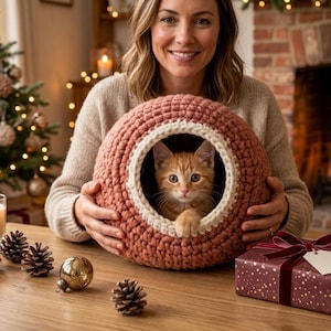 Puede incluir: Una cama para gatos de ganchillo en color rosa empolvado con un ribete color crema, que alberga un gatito jengibre. La cama para gatos está sostenida por una persona. Decoraciones navideñas, incluyendo un árbol, adornos y una caja de regalo, están en el fondo.