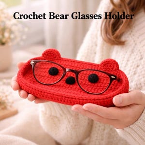 May include: A red crochet bear glasses holder with black accents and two small ears. The glasses holder is holding a pair of eyeglasses with a dark brown frame. The text "Crochet Bear Glasses Holder" is at the top of the image.