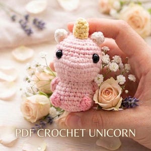 May include: A small, pink crocheted unicorn with a yellow horn, black eyes, and white ears. The unicorn is surrounded by flowers and the text "PDF CROCHET UNICORN".