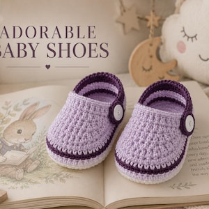 May include: A pair of handmade crochet baby shoes in light purple and dark purple yarn. The shoes feature a rounded toe, a dark purple striped trim, and a single white button on the side. The text ADORABLE BABY SHOES is visible above the shoes.