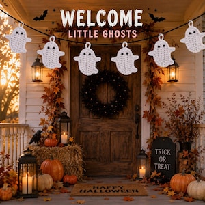 May include: A string of six white crocheted ghost ornaments hanging on a black cord. The ghosts have black eyes and are suspended in front of a wooden door decorated with autumn leaves, pumpkins, lanterns, and a sign reading WELCOME LITTLE GHOSTS.