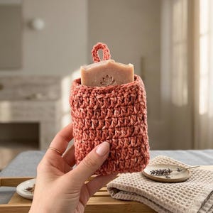 May include: A coral-colored, crocheted soap holder with a loop for hanging. The holder contains a bar of soap with a lotus flower design. The soap holder is displayed on a wooden tray with a small ceramic dish and a folded waffle-weave towel.
