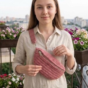 May include: A dusty rose crochet fanny pack with a matching strap and silver zipper. The bag is worn across the body. The bag is made of a textured, woven material.