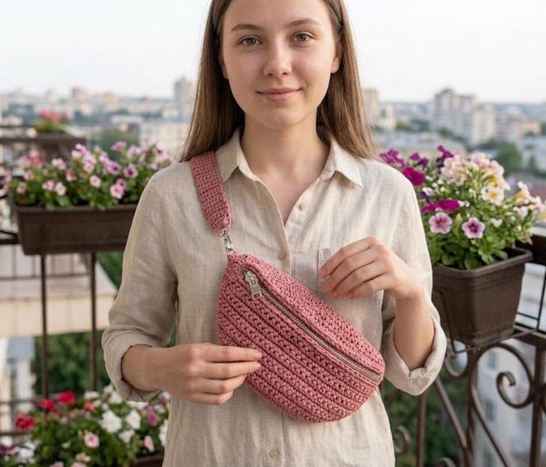 May include: A dusty rose crochet fanny pack with a matching strap and silver zipper. The bag is worn across the body. The bag is made of a textured, woven material.