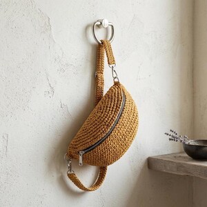 May include: A mustard-colored, crocheted fanny pack with a black zipper and silver hardware hangs from a silver hook on a textured white wall. The bag has a curved shape and a strap that can be adjusted. The fanny pack is a stylish accessory.