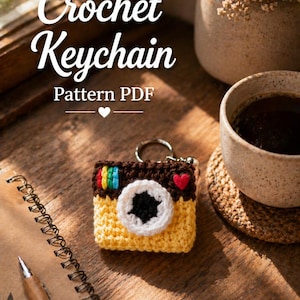 Puede incluir: Un llavero de cámara de ganchillo en amarillo, marrón, blanco y colores arcoíris. El llavero tiene una lente negra y un detalle de corazón rojo. El texto "Crochet Keychain Pattern PDF" es visible en la imagen.