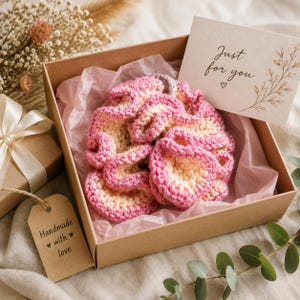 May include: A round, ruffled crochet accessory in shades of pink and cream, nestled in a brown cardboard gift box lined with pink tissue paper. A white card with the text Just for you and a heart icon is included, alongside a brown paper tag.