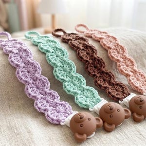 Puede incluir: Cuatro clips de chupete de ganchillo en colores pastel: lavanda, verde menta, marrón y melocotón. Cada clip tiene un extremo en bucle y un accesorio de silicona con forma de oso. Los clips están colocados sobre una superficie de tono neutro.