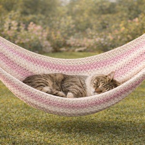 Può includere: Un gatto tigrato dorme pacificamente in un'amaca all'uncinetto. L'amaca, con un motivo a righe rosa e bianche, è sospesa tra due pali di legno. La scena è ambientata all'aperto, in un'area erbosa.