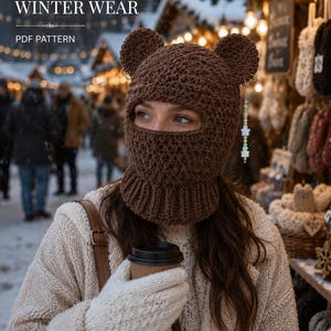 May include: A brown crocheted balaclava featuring two rounded bear ears on top and a ribbed neck collar. A silver hoop earring with a dangling beaded charm featuring star-shaped beads hangs from the left ear. Text reads HANDMADE WINTER WEAR PDF PATTERN.