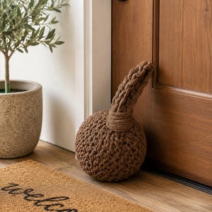May include: A spherical doorstop crafted from thick, brown woven rope. The object features a dense, textured knot pattern with a sturdy handle loop extending from the top, designed to hold a door in place with its substantial, rounded shape.