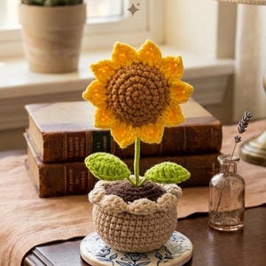 Puede incluir: Un girasol de ganchillo hecho a mano en una maceta pequeña. El girasol tiene pétalos amarillos y un centro marrón. La maceta es beige y se asienta sobre un posavasos decorativo. Las hojas son verdes. El tema general es la decoración del hogar.