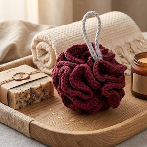 May include: A wooden tray holds a burgundy knitted bath pouf with a white loop, a bar of soap wrapped in brown paper, a rolled beige towel, and a lit candle in a glass jar. The items create a spa-like atmosphere.